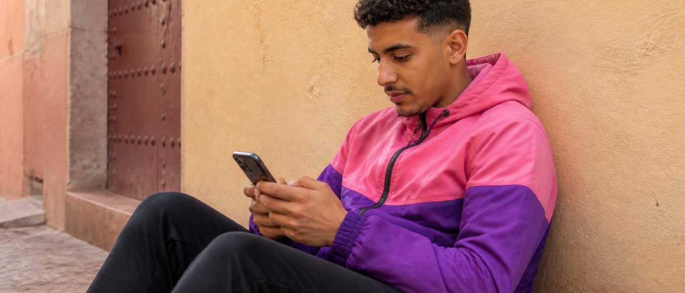 North African young man sitting alone with his phone against a wall